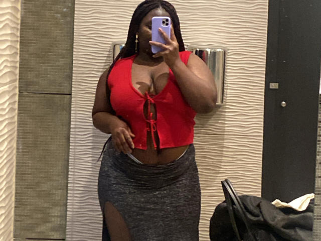 Dilay with dark skin and long braids, wearing a red crop top with a lace-up front, showing her tits, and a gray skirt with a thigh slit, taking a selfie.