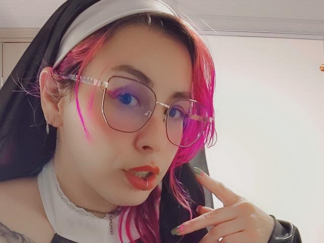 NatashaLau with pink and black hair, nun habit, glasses, red lipstick, and green nails, showing off pierced tongue and finger to mouth. Her tits are partially visible.
