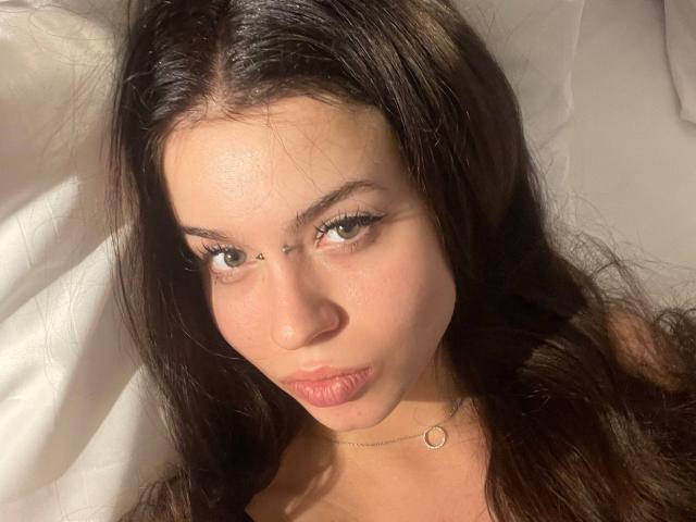BrandyCrow has long dark hair, green eyes, and a small metal chain on her forehead. She's wearing a thin gold necklace, and her lips are slightly puckered.