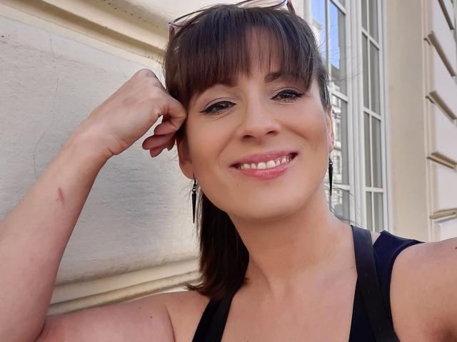 LaLeticia-hot with brown hair, bangs, and sunglasses on head, smiling, black tank top, pink lipstick, small scar on arm, standing against white wall, window on right.