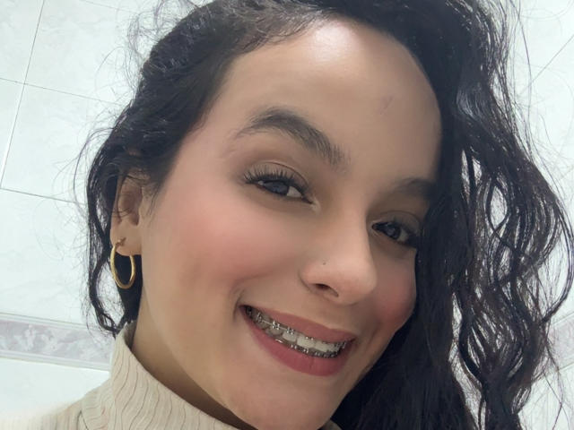 MaiaPark has a cute smile with metal braces, wearing a white turtleneck, gold hoop earrings, and wavy dark hair. Her brown eyes are slightly upturned.