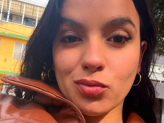 MaiaPark has brown skin, dark eyebrows, long eyelashes, and pink lips. She wears a brown leather jacket, hoop earrings, and has a small nose piercing. Her expression is neutral.