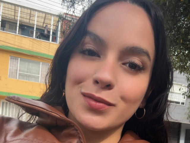 MaiaPark's face, brown leather jacket, hoop earrings, long dark hair, slightly smiling, brown eyes, subtle makeup, soft pink lips, tree and yellow building in background, daytime.