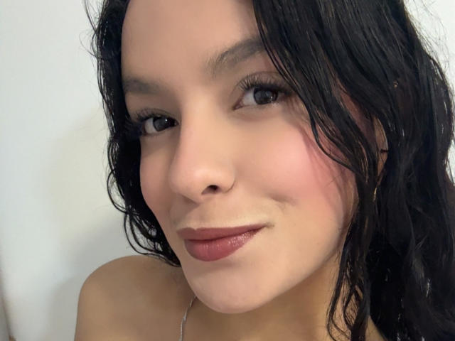 MaiaPark has wavy black hair, light skin, and is smiling with red lipstick. Her small tits are slightly visible. She's wearing a silver necklace.