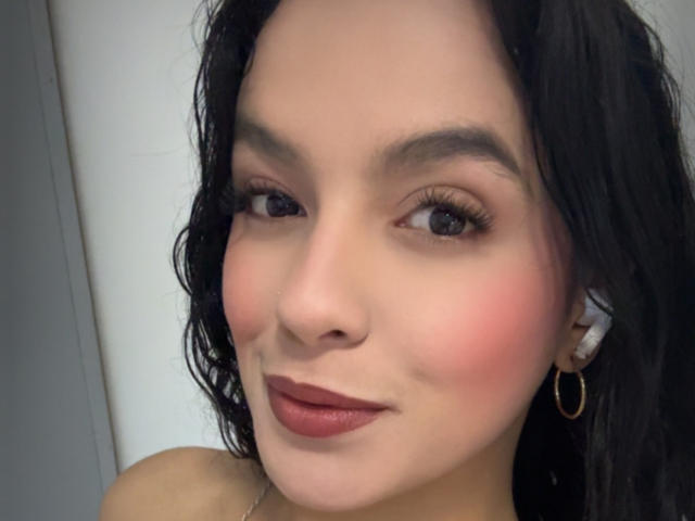 MaiaPark has fair skin, black wavy hair, brown eyes, and pink blush. She's wearing dark red lipstick and gold hoop earrings. Her face is slightly tilted to the right.