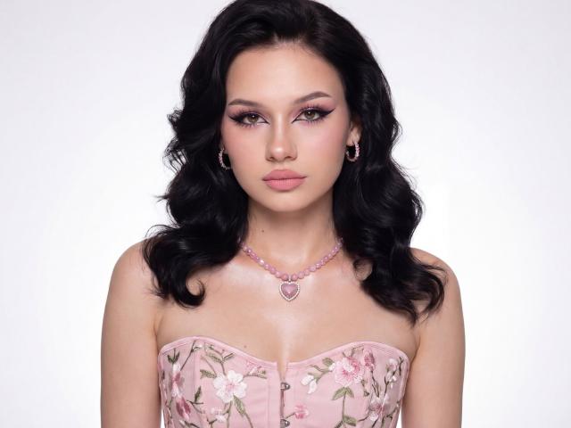 AlicePertim with black wavy hair, pink floral strapless dress, pink necklace with heart pendant, and pink makeup, looking straight at the camera with a neutral expression.