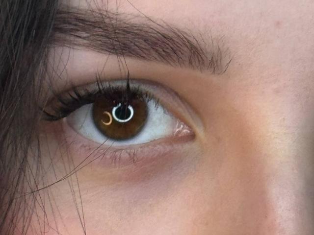 MeganCarlson's brown eye with a small silver ring piercing on the upper eyelid, thick dark eyebrows, and long black eyelashes, showing a slightly pinkish skin tone.