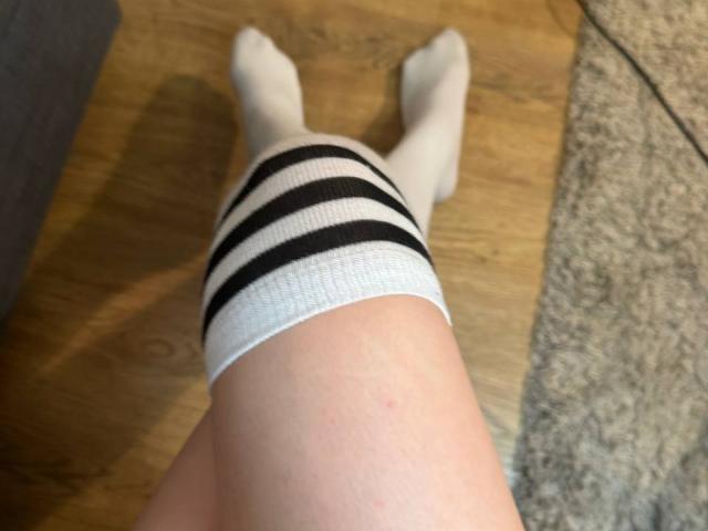 MegganCarlson's legs wearing white knee socks with black stripes, lying on a wooden floor with a gray carpet on the right. Her pale skin is visible.