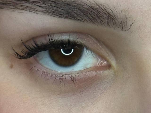 MegganCarlson's brown eye with long black eyelashes, a small beauty mark below, and thick dark eyebrows. Her skin is light with a slight pinkish hue.