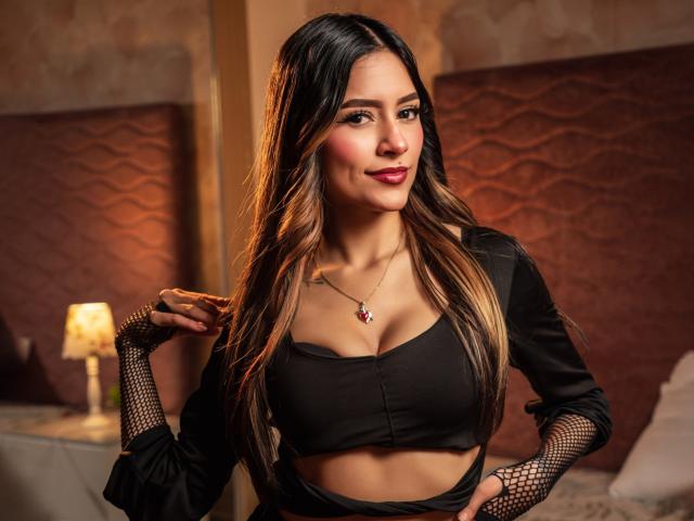 AfroditaWild has long, wavy brown hair, wearing a black crop top that shows her tits, fishnet gloves, and a red pendant necklace, standing in a warmly lit room.