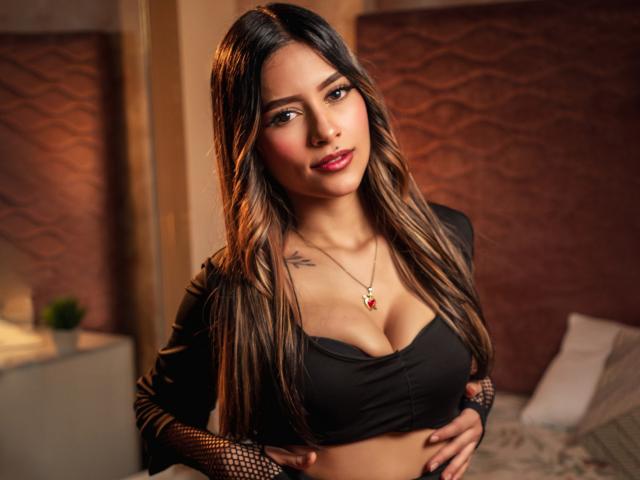 AfroditaWild with long, wavy brown hair, black crop top, and fishnet sleeves, has a tattoo on her right shoulder, showing off her boobs and a red necklace.