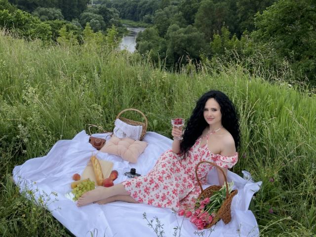 VictoriaAlarie with black curly hair, floral dress, bare shoulders, holding wine glass, sitting on white blanket, surrounded by picnic basket, bread, grapes, and basket with roses.