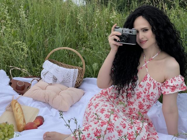 VictoriaAlarie with long black curly hair, wearing a pink floral dress, sitting on a white blanket, holding a vintage camera, with a wicker basket, grapes, bread, and peaches.