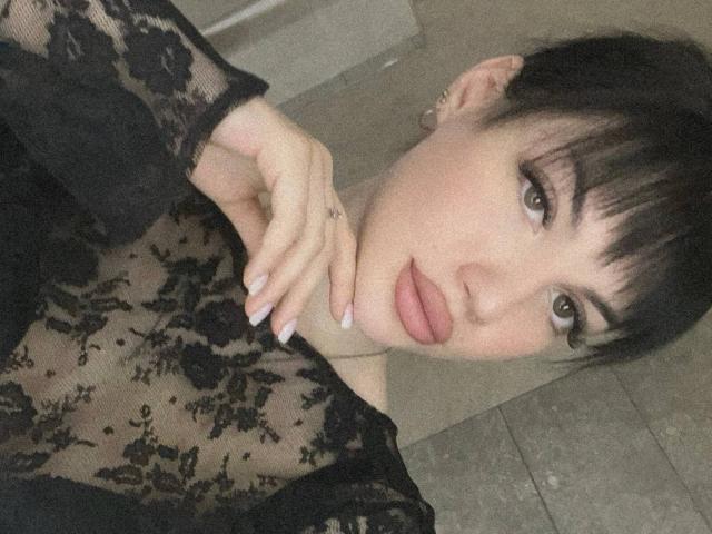 Natalia Liukko con un vestido de encaje negro, piel pálida, cabello oscuro recogido en un moño, maquillada, tocándose el cuello, mostrando sus pechos a través de una tela transparente, en un selfie..