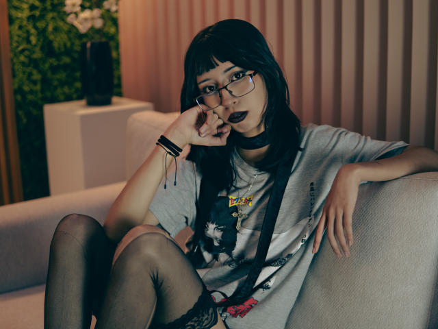 YumeLee with black hair, glasses, and dark lipstick, wearing a gray shirt, black choker, and thigh-high stockings, sitting on a sofa, hand near mouth, looking at camera.