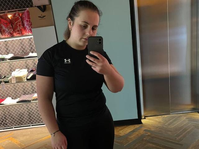 DariaCalm with brown hair in a ponytail, black Under Armour shirt, and black pants, taking a mirror selfie with her iPhone in a store with wooden floors.