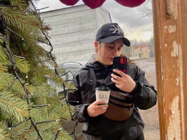 DariaCalm with a black cap, holding a coffee cup in one hand and a phone in the other, standing in front of a decorated Christmas tree.