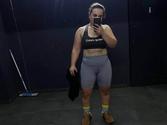 DariaCalm stands in a dimly lit room, wearing a black sports bra and gray shorts, holding a black shirt, with tan boots and yellow socks, taking a mirror selfie.