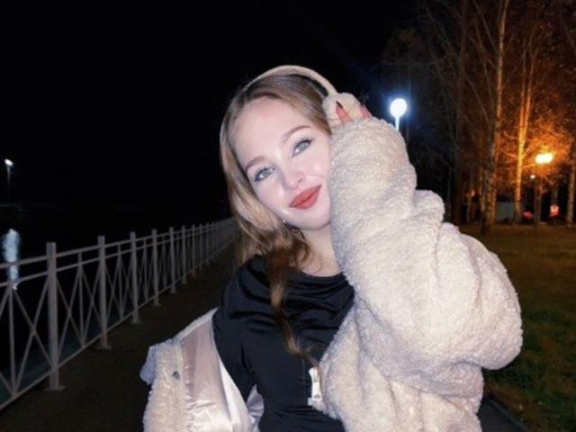 AileneArimas has blonde hair, blue eyes, and wears a fluffy beige jacket with a black top, holding a pink bag, and a beige headband, smiling at night.