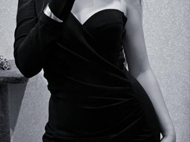 AileneArimas in a tight, black, strapless dress, showing her bare shoulders and upper chest, with a black sleeve on her right arm, holding a dark object.