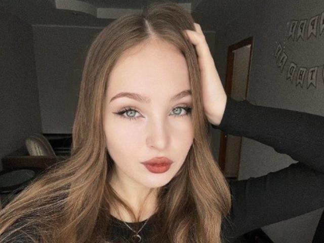 AileneArimas has light green eyes, long brown hair, and wears dark lipstick with black eyeliner. She's touching her hair with one hand, wearing a black top.