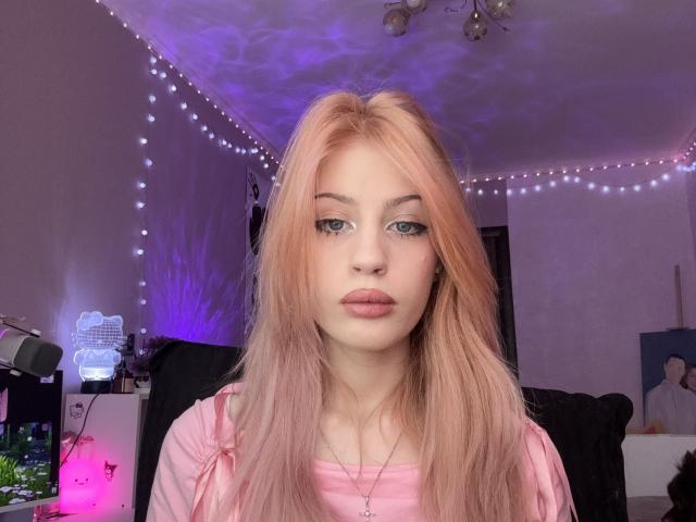 PeiMrozoski has pinkish-blonde hair, blue eyes, and pink lips. She's wearing a pink shirt, looking at the camera with a neutral expression. Room is dimly lit with string lights.