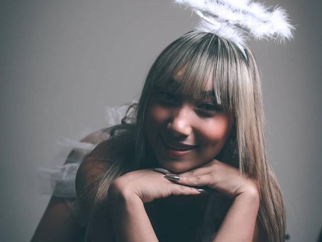 EvaGreeny met blond haar en pony, lachend, leunend op haar handen, gekleed in witte engel vleugels en een halo, grijze achtergrond, zachte belichting, ziet er schattig en aantrekkelijk uit..