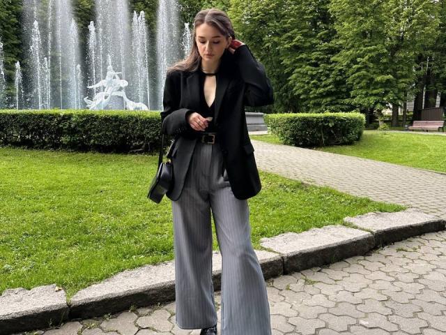 CyrenaLuxe with gray pants, black blazer, and black handbag, standing on a hexagon-patterned path, adjusting her hair, with a water fountain in the green background.