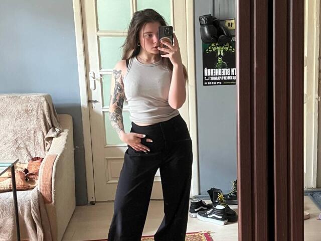 Mettis with a tattooed arm, wearing a white tank top and black pants, taking a mirror selfie, hand on her butt, in a casual room, showing her tits.
