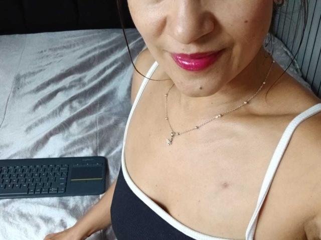 SolaraDubois with pink lipstick, black tank top, white straps, small boob, nipple slightly visible, collarbone necklace, black keyboard, bed, light skin, short brown hair, subtle smile, slight bruise.