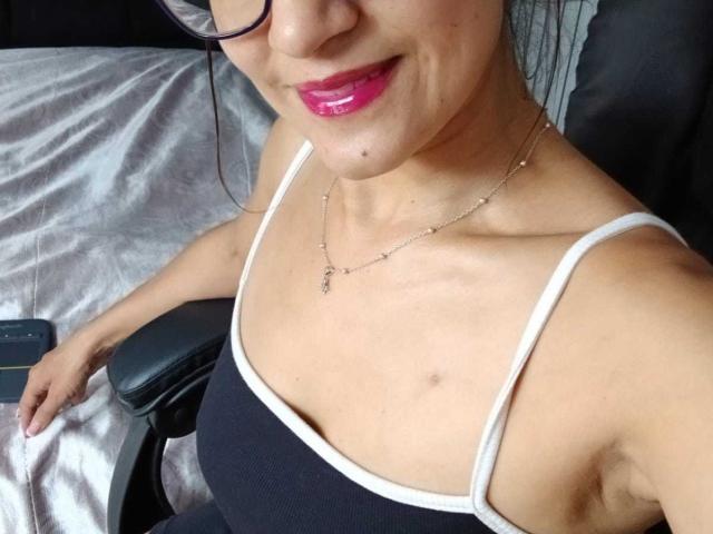 SolaraDubois with pink lipstick, black tank top, white straps, necklace, glasses, visible armpit, small breast, light skin, sitting in chair, holding phone.