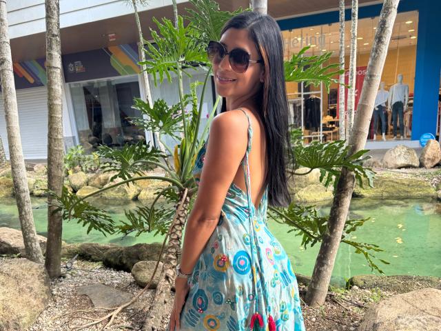 Chlarise, avec ses longs cheveux noirs, portant des lunettes de soleil et une robe bleue ornée de cercles colorés, souriant à l'appareil photo, se tenant près d'un petit étang vert avec des rochers et des arbres..