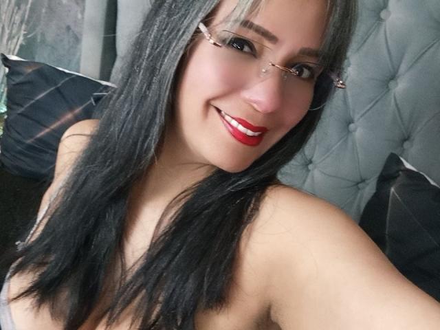 Laila Castillo, avec de longs cheveux noirs et blonds, des lunettes, du rouge à lèvres rouge, souriante, montrant ses seins, portant un débardeur gris, assise sur une tête de lit grise capitonnée..