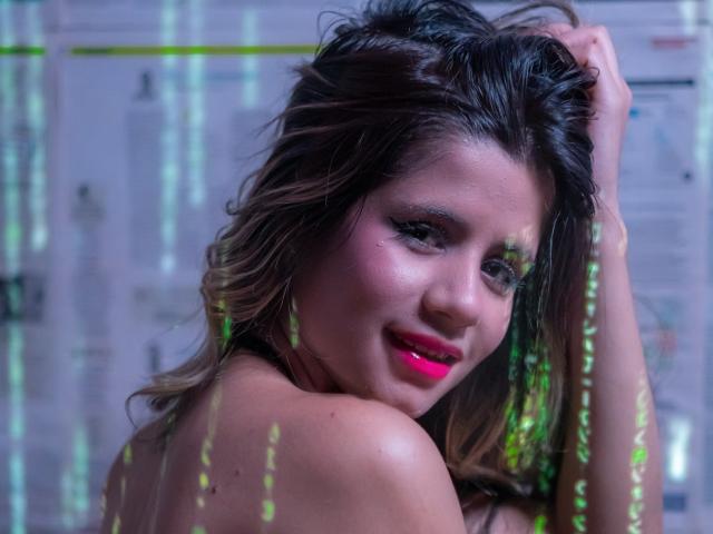 SheylaSpooner with pink lipstick, dark wavy hair, and a smile, touching her head with one arm, green light reflections on her skin, topless, looking over her shoulder.