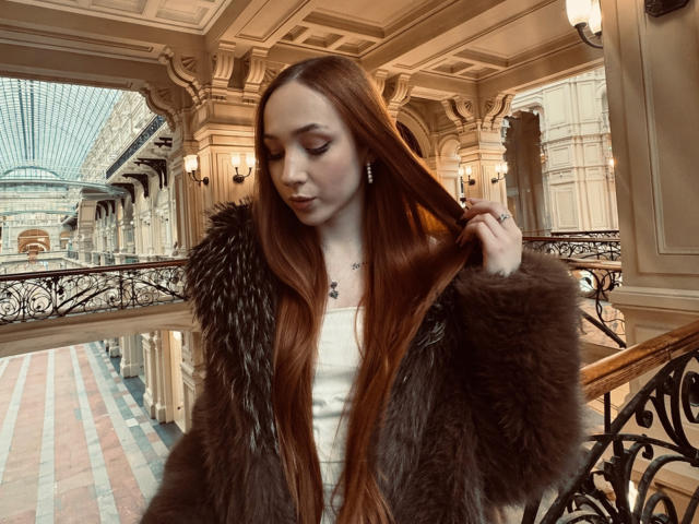 Alona Hadcock, con cabello largo y pelirrojo, vestida con una chaqueta de piel marrón, sujetándose el cabello, mirando hacia abajo, en un entorno interior elegante y ornamentado con un techo de cristal..