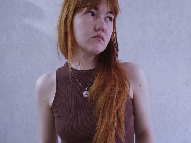 SheritaChieng has red hair, a nose ring, and a brown sleeveless top, with a silver necklace and pouty expression, showing her small tits and freckles.