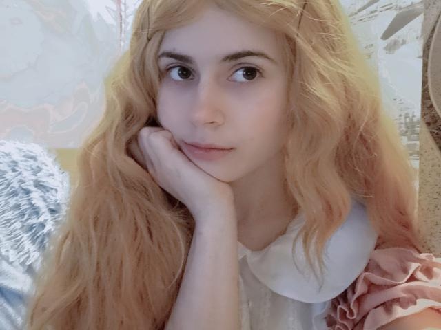 Mireille Spotwood a de longs cheveux blonds ondulés, un visage pâle, et repose son menton sur sa main avec une expression douce et neutre. Elle porte une robe rose et blanche..