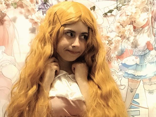 MireilleSpotwood with long, wavy, orange wig, smiling, hands near neck, wearing pink and white dress, pale skin, subtle makeup, X-chip in hair, anime-style background, cute.