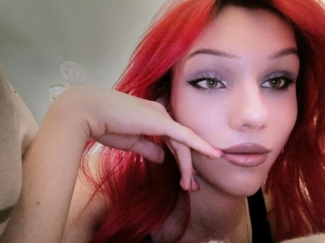 KiranaHayes with bright red hair, pouty lips, and gray eyeshadow, touching her chin with a finger, wearing black top, showing subtle makeup and clear skin.