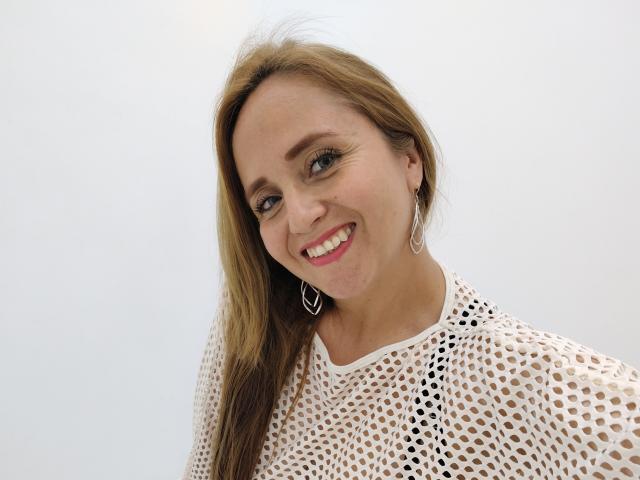 CamilaLux has long blonde hair, blue eyes, and a smile showing pink lipstick. She's wearing a white mesh top and silver heart-shaped earrings. Her tits are slightly visible.