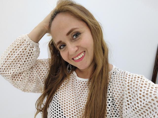 CamilaLux, com cabelos castanhos longos, sorrindo, vestindo uma blusa branca de tela, com a mão no cabelo, brincos, mamilos visíveis e batom rosa..
