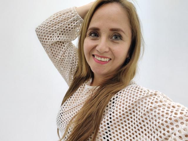 CamilaLux mit langen, braunen Haaren, lächelnd, trägt ein weißes, durchsichtiges Oberteil, hat eine Hand in den Haaren und ihre Brüste sind durch die Löcher hindurch sichtbar, sie trägt pinken Lippenstift, hat blaue Augen, helle Haut und ein süßes Gesicht..