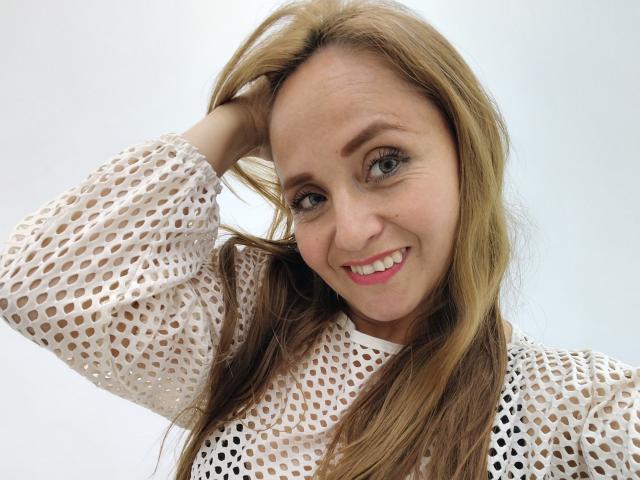 CamilaLux mit blonden Haaren, lächelnd, trägt ein weißes, durchsichtiges Oberteil, hat eine Hand im Haar, zeigt kleine Brüste, trägt pinken Lippenstift und hat blaue Augen, vor einem schlichten Hintergrund..