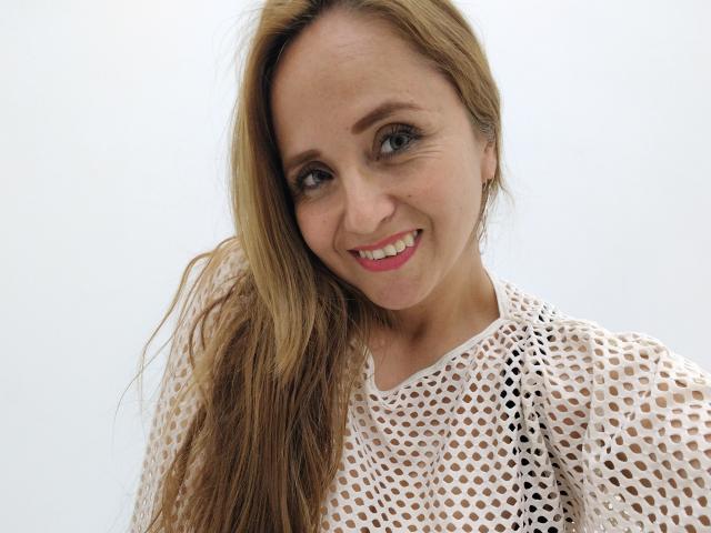 CamilaLux mit langen, blonden Haaren, lächelnd, trägt ein weißes, durchsichtiges Oberteil, das ihre Brüste zeigt. Sie hat rosafarbenen Lippenstift, grüne Augen und blickt in die Kamera..