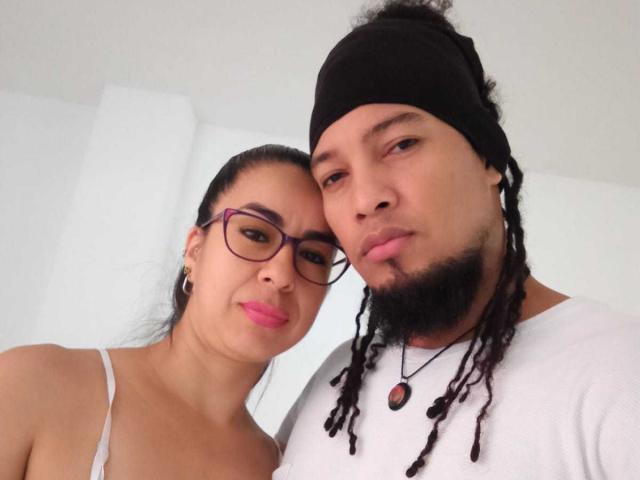 Eine Frau namens MinervaHot mit Brille, rosa Lippenstift und kleinen Brüsten, steht neben einem Mann mit schwarzer Mütze, weißem Hemd und Dreadlocks, beide schauen in die Kamera..