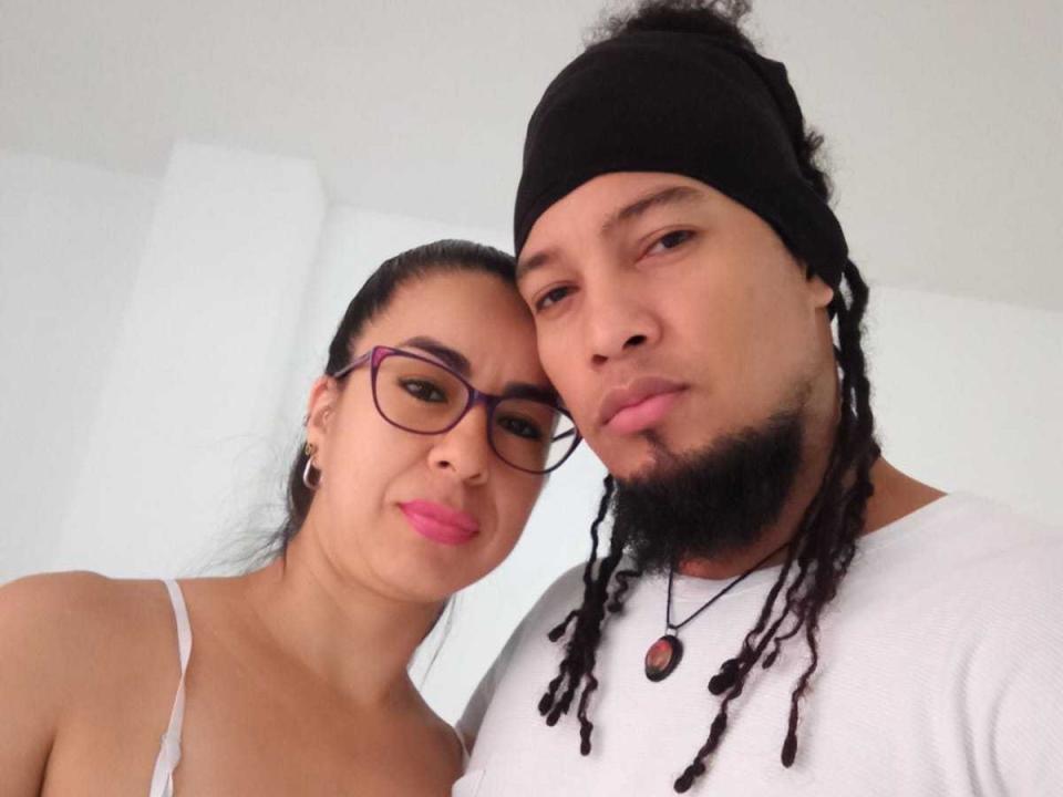 Minerva, con gafas, lápiz labial rosa y senos pequeños, junto a un hombre con un gorro negro, camiseta blanca y rastas, ambos mirando a la cámara..