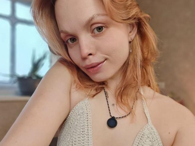 Sinteas has pale skin, green eyes, and red hair, wearing a crocheted top, black necklace with a round pendant, and a subtle smile, showing her small tits.