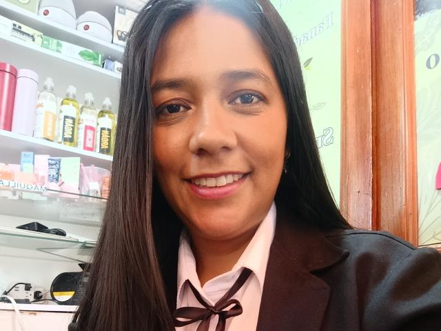 PauletteLoveX with long brown hair, tan skin, and pink lipstick, smiling, wearing a dark brown blazer over a white shirt with a black ribbon. Shelves with products in background.