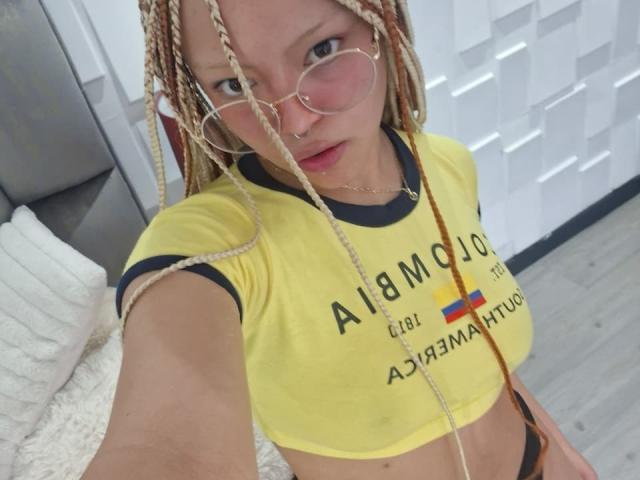 KeiryGinger with blonde braids, round glasses, nose ring, yellow crop top, showing her small tits, and black panties, taking a selfie in a bedroom.