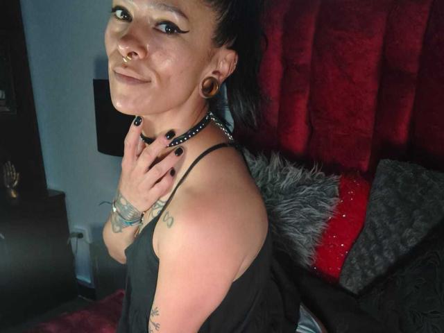 AmelieBlake with black hair in a ponytail, black eyeliner, nose ring, black choker, black nails, tattoos, black top, hand on neck, smiling, red curtain background.